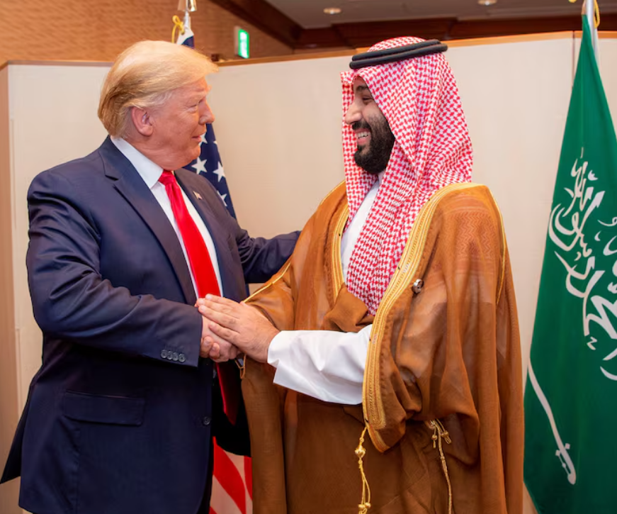 Trump and MBS shake hands in front of the U.S. and Saudi Arabian flags.